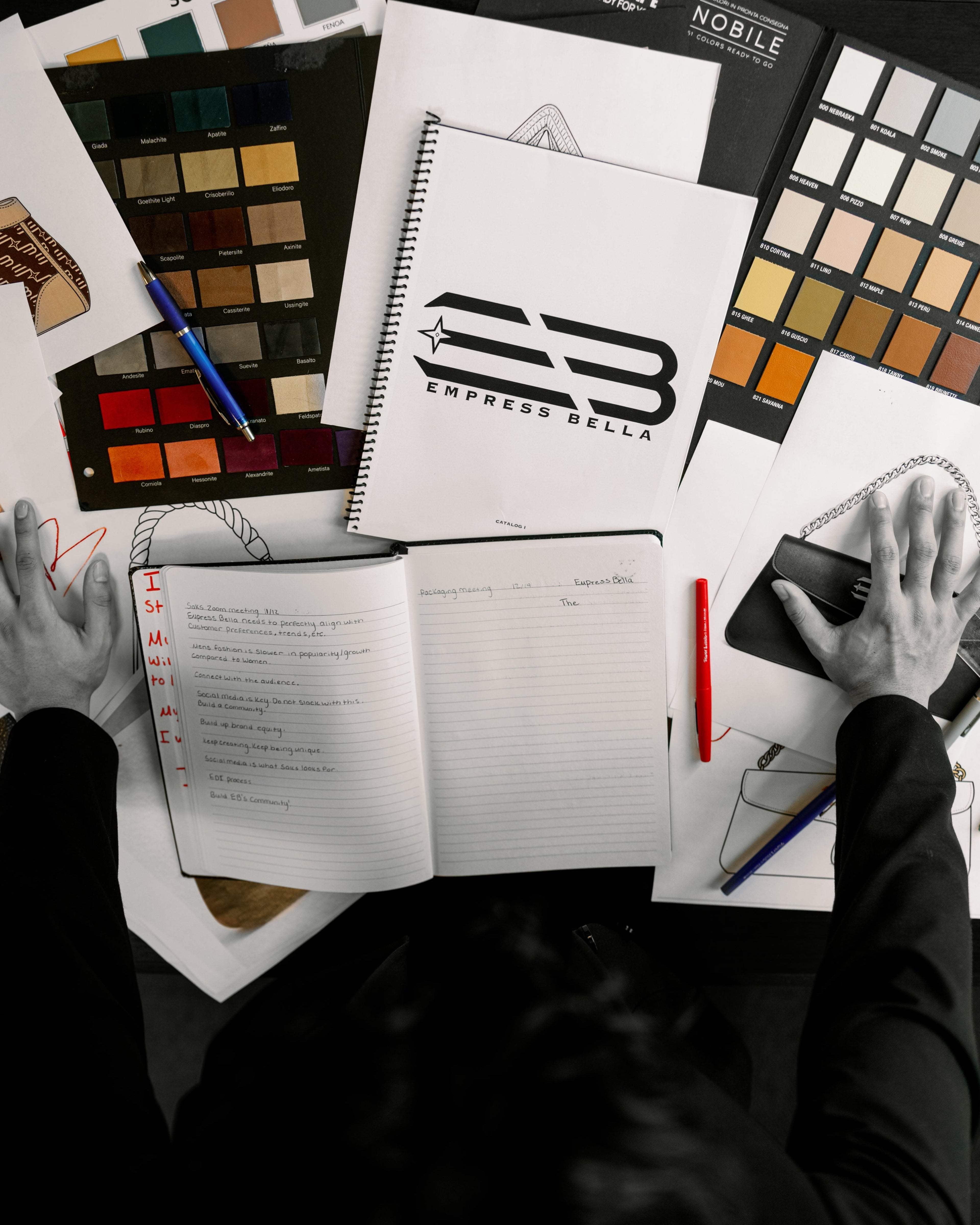 Overhead view of a designer's desk with sketches, color swatches, a notebook with the logo 'EB Impress Bella', and a phone being held by hands in a black long-sleeve top.